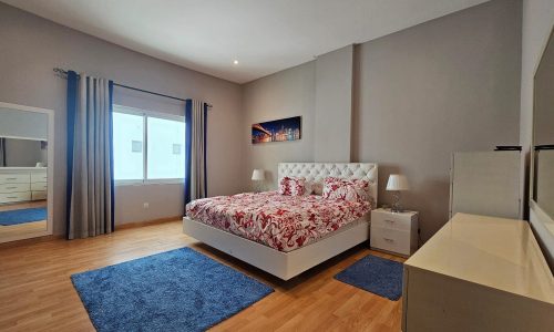 Modern bedroom with hardwood floor, double bed with red and white bedding, two nightstands with lamps, a dresser, a large mirror, and a window with gray curtains. Two blue rugs are on the floor.