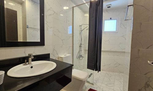 A bathroom featuring a sink with a mirror, a toilet, and a glass-enclosed shower area with a curtain. The walls and floor are tiled with white and gray patterns, and a small window is near the shower.