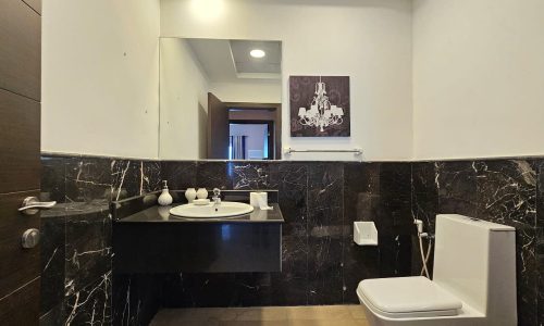 A modern bathroom features a wall-mounted sink, a toilet, a mirror, black marble wall tiles, and a chandelier artwork on the wall. A door is partially open, revealing another room in the background.