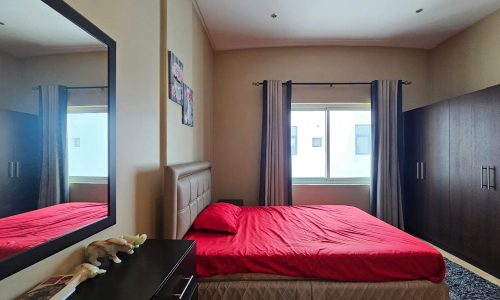 A bedroom with a red bedspread, dark wooden wardrobes, a window with curtains, and a mirror reflecting the room. There is a bedside table with items on it and framed pictures on the wall.