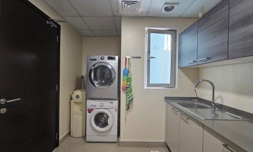 A laundry room with a stackable washer and dryer, a sink with a faucet, overhead cabinets, and a window. Cleaning supplies are also present.