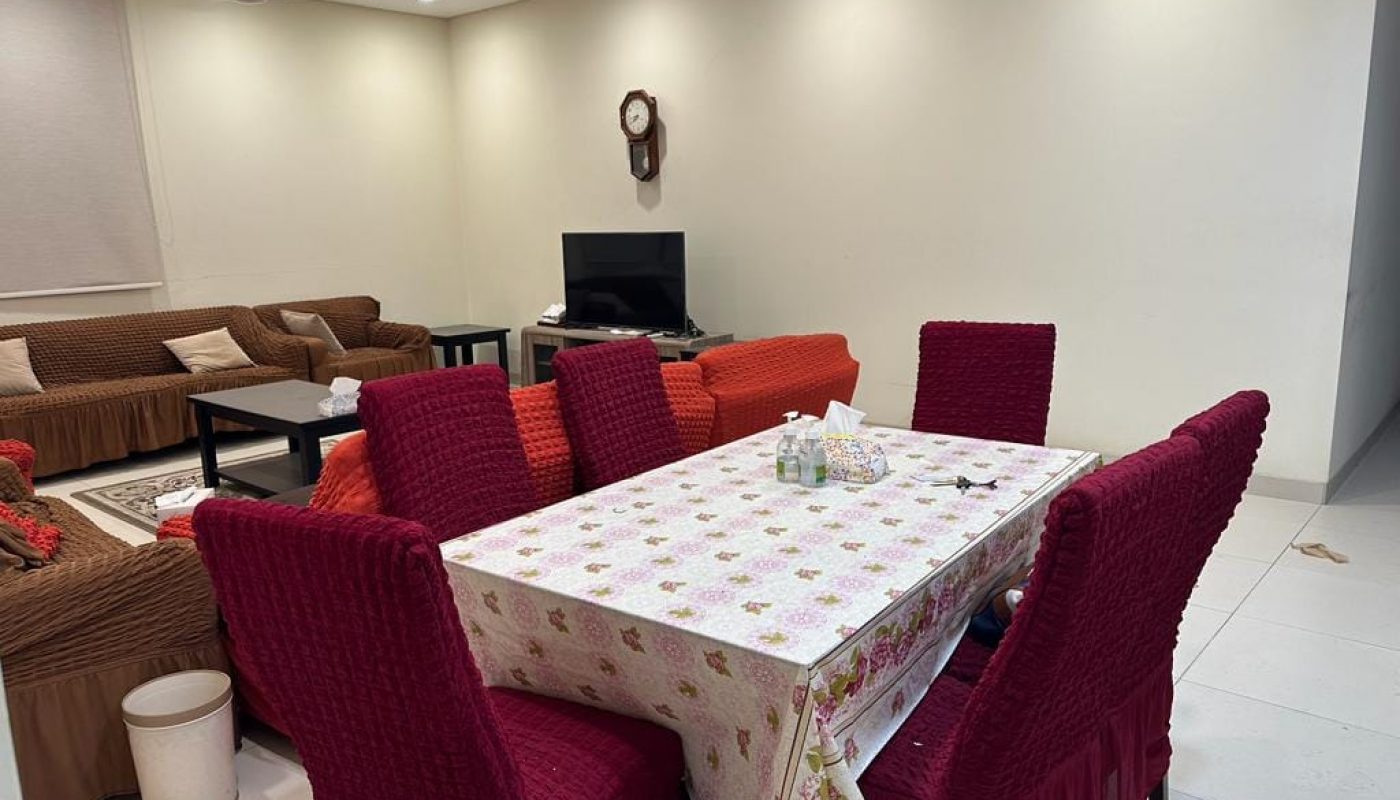 Dining room with red chairs and a floral tablecloth. In the background, a living room with brown sofas and a TV. Walls are light, and there's a wall clock above the TV.