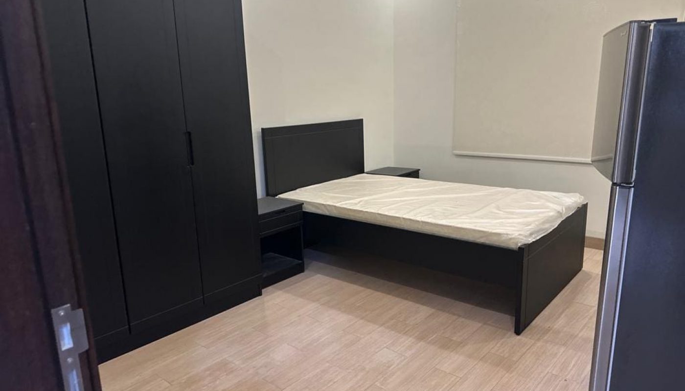 Room with a dark wardrobe, bed with a white sheet, a bedside table, light wood floor, and a refrigerator in the corner.