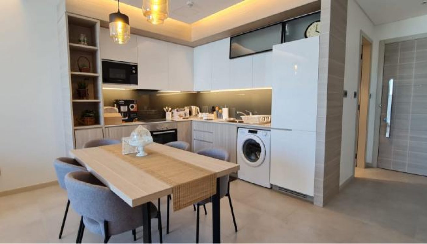 Modern kitchen and dining area with a wooden table, four chairs, built-in appliances, washing machine, and shelves with decor, illuminated by pendant lights.