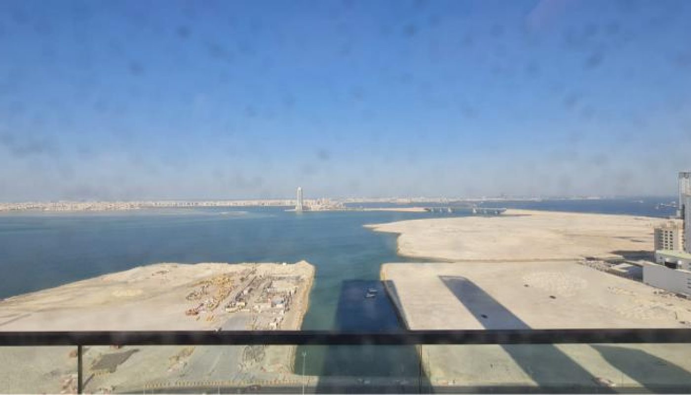 View from a window overlooking a construction site by the waterfront with a clear blue sky, city buildings, and distant shoreline visible across the water.