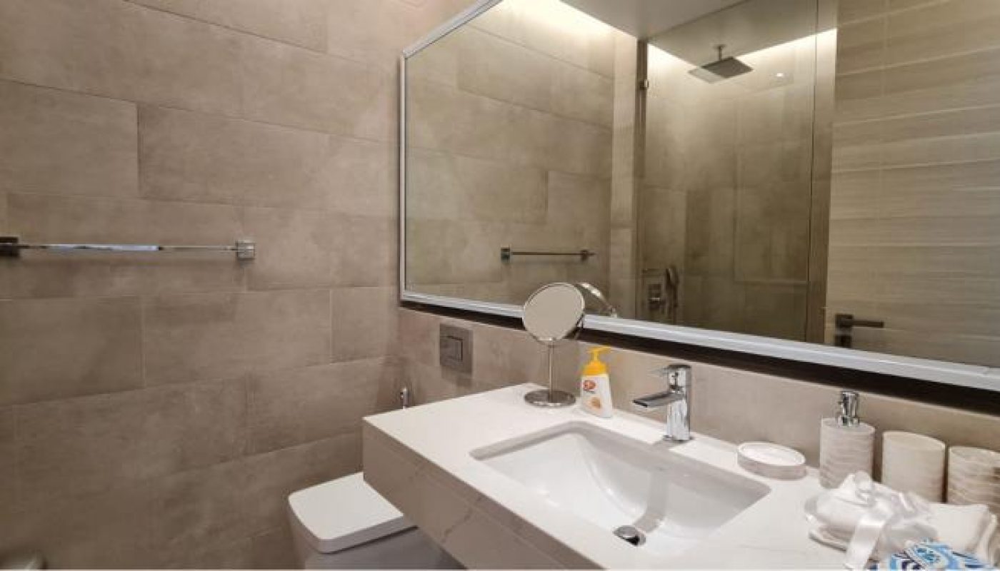 Modern bathroom with beige tile walls, large mirror, white sink, toiletries, towel rack, and a walk-in shower with a rainfall showerhead reflected in the mirror.