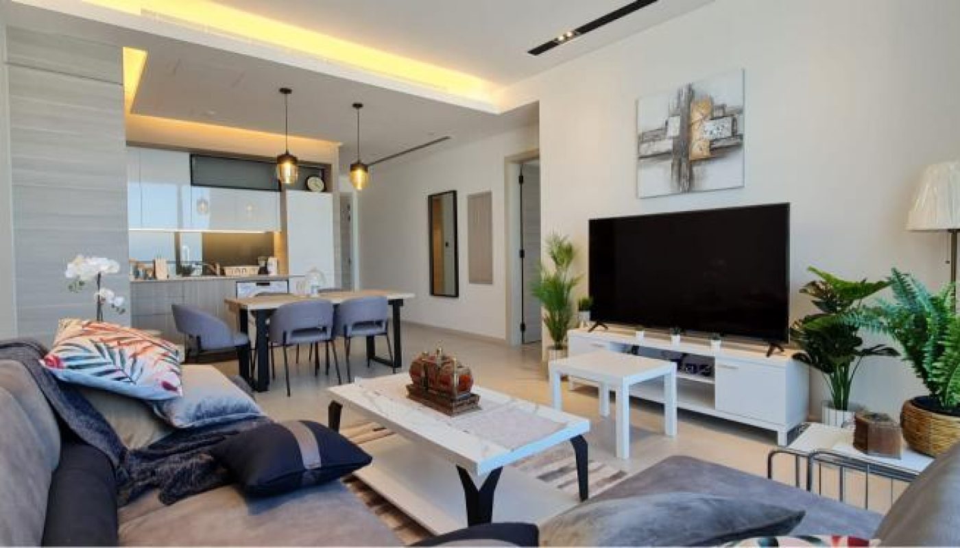 Modern living room with a gray sofa, white coffee table, large TV, indoor plants, dining area, and an open kitchen in the background.