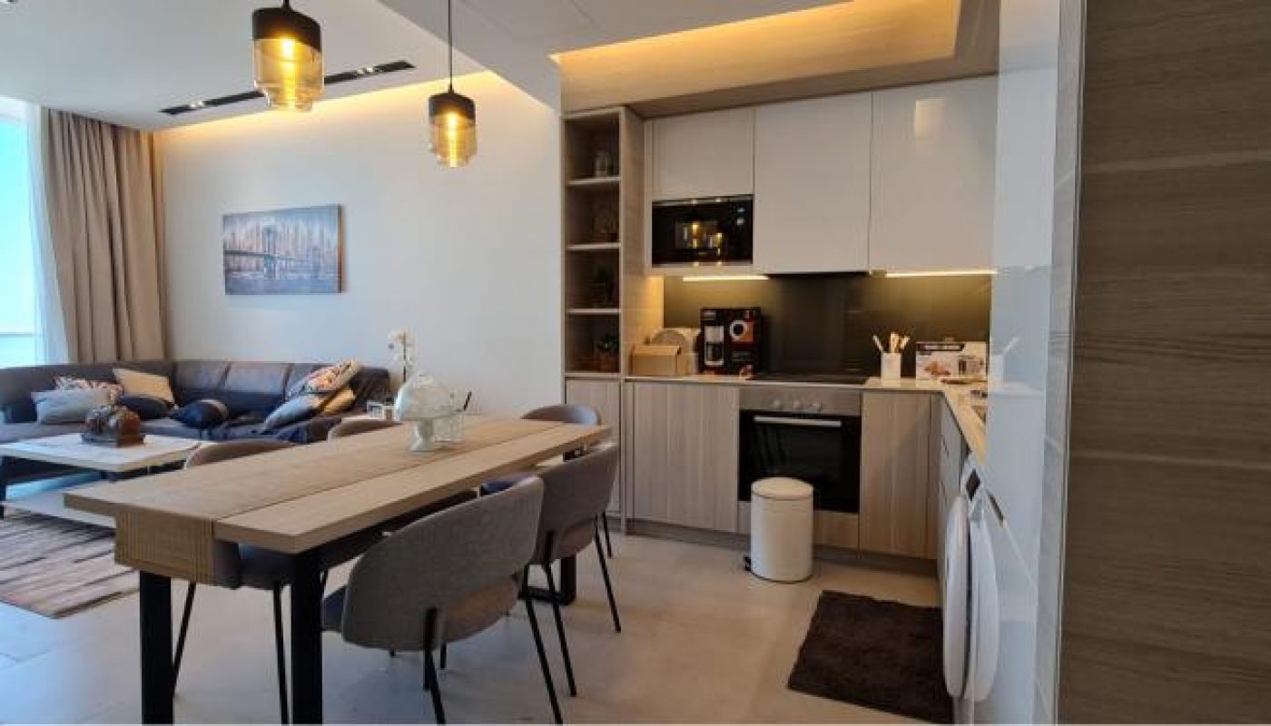 Modern open-plan kitchen and living area with a dining table, sofa, large window, and ceiling lights, featuring neutral colors and contemporary furnishings.