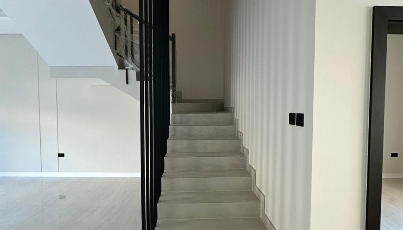 A modern, minimalistic staircase with gray steps and a black handrail ascends beside a white wall in a spacious, light-filled room with shiny tile flooring.