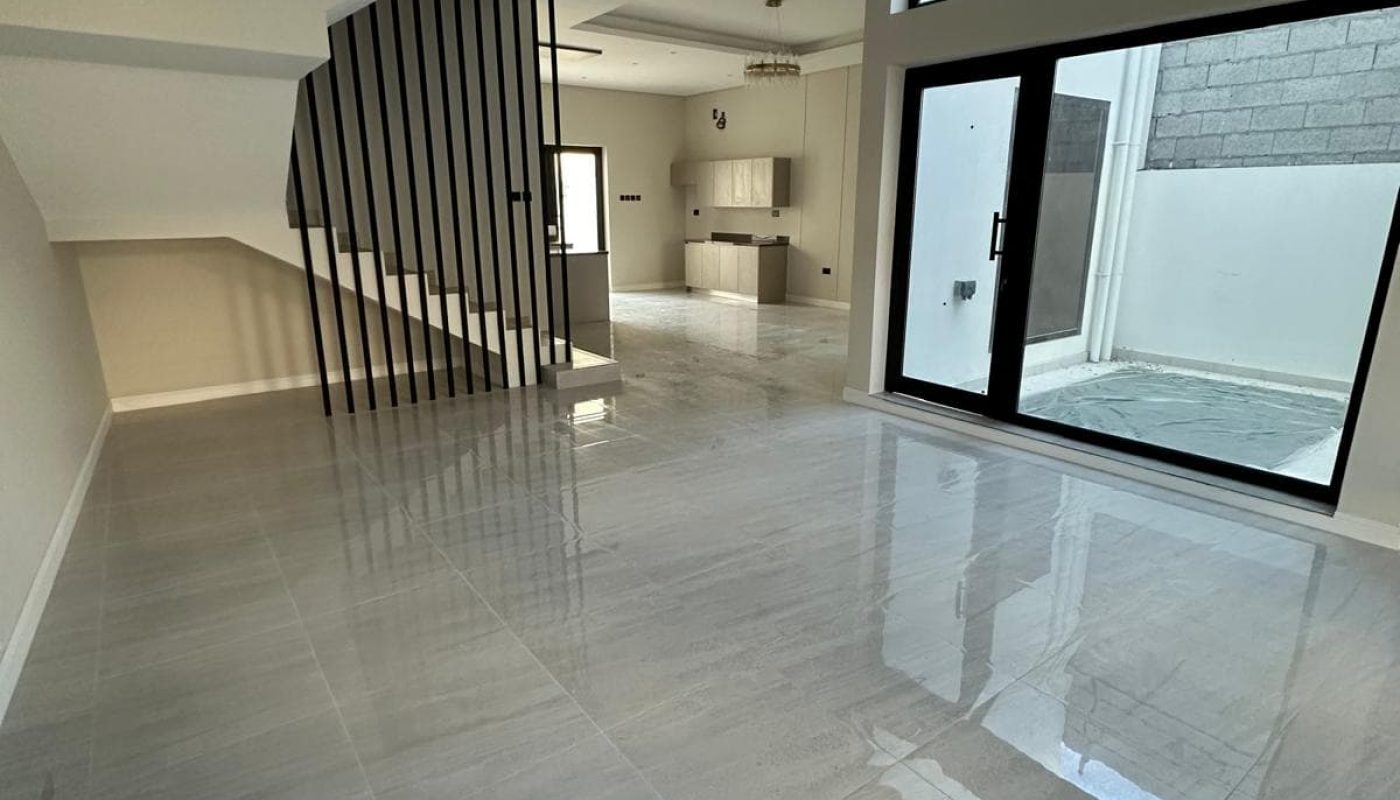 Spacious empty room with glossy tiled floors, large windows, a modern chandelier, and a staircase with black railings leading to an upper level.