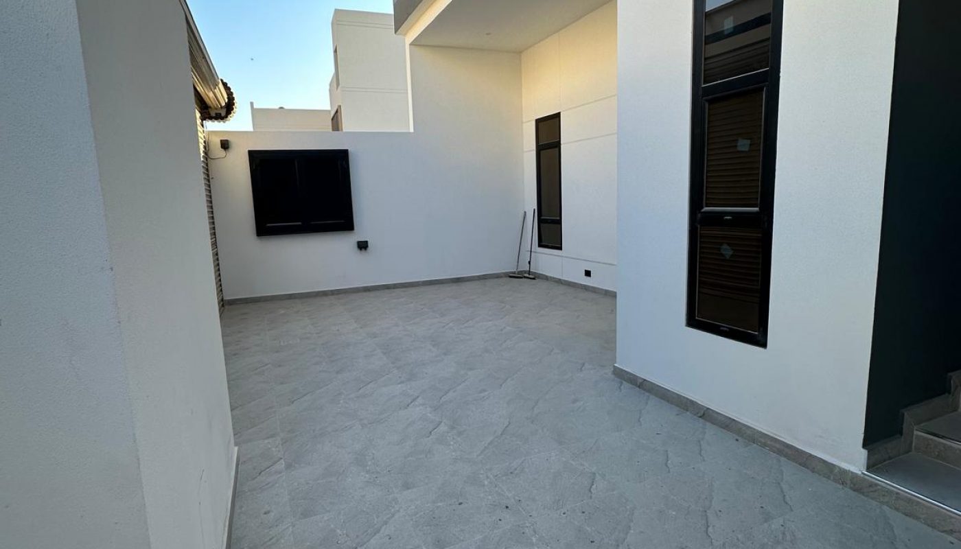 Outdoor area with gray tile flooring next to a modern building facade. The facade features windows and a wood panel accent. Sky is clear. Stairs are visible on the right.