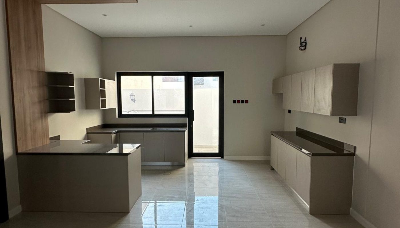 A modern kitchen with neutral cabinetry, large floor tiles, a central island, and a window in the background.