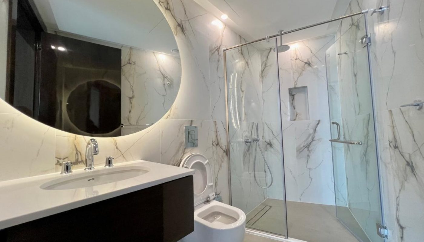Modern bathroom with a double sink, illuminated round mirror, open toilet, and glass-enclosed shower; marble-patterned walls and neutral flooring.