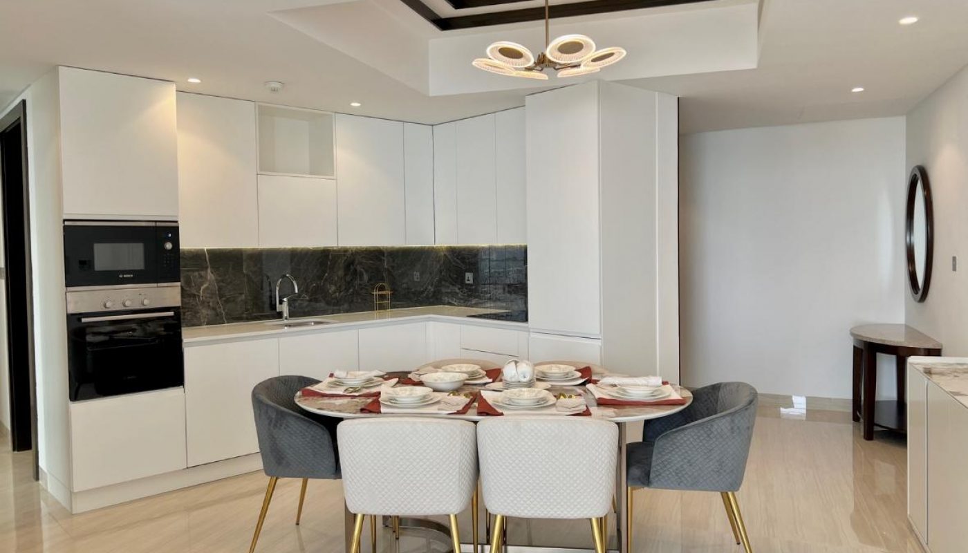 Modern kitchen with white cabinets, a black backsplash, built-in appliances, and a dining table set for six with gray and white chairs. Round light fixture hangs above the table.
