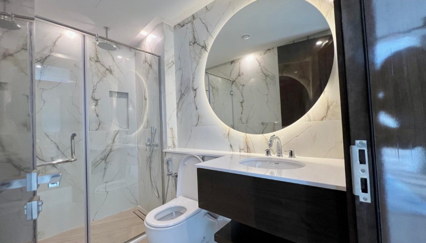 Modern bathroom with marble-pattern walls, glass-enclosed shower, wall-mounted toilet, floating vanity with sink, and a large round backlit mirror.