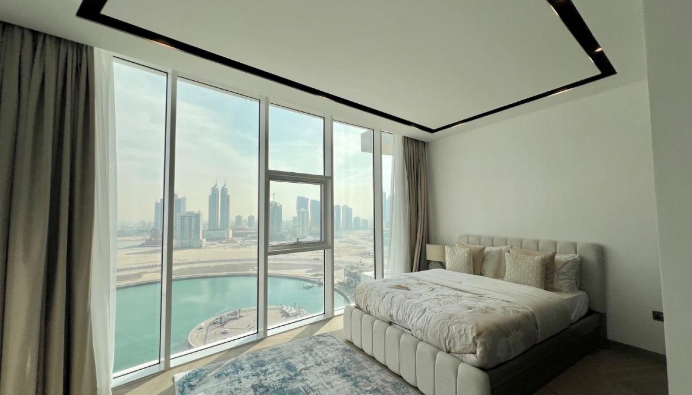 A modern bedroom with a large bed, floor-to-ceiling windows, city skyline and water views, neutral decor, and a patterned area rug.