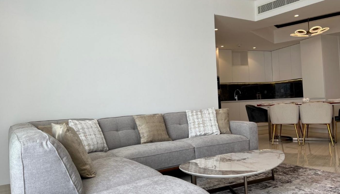A modern living room with a grey sectional sofa, marble coffee table, and a view of an open kitchen and dining area with gold-accented chairs.