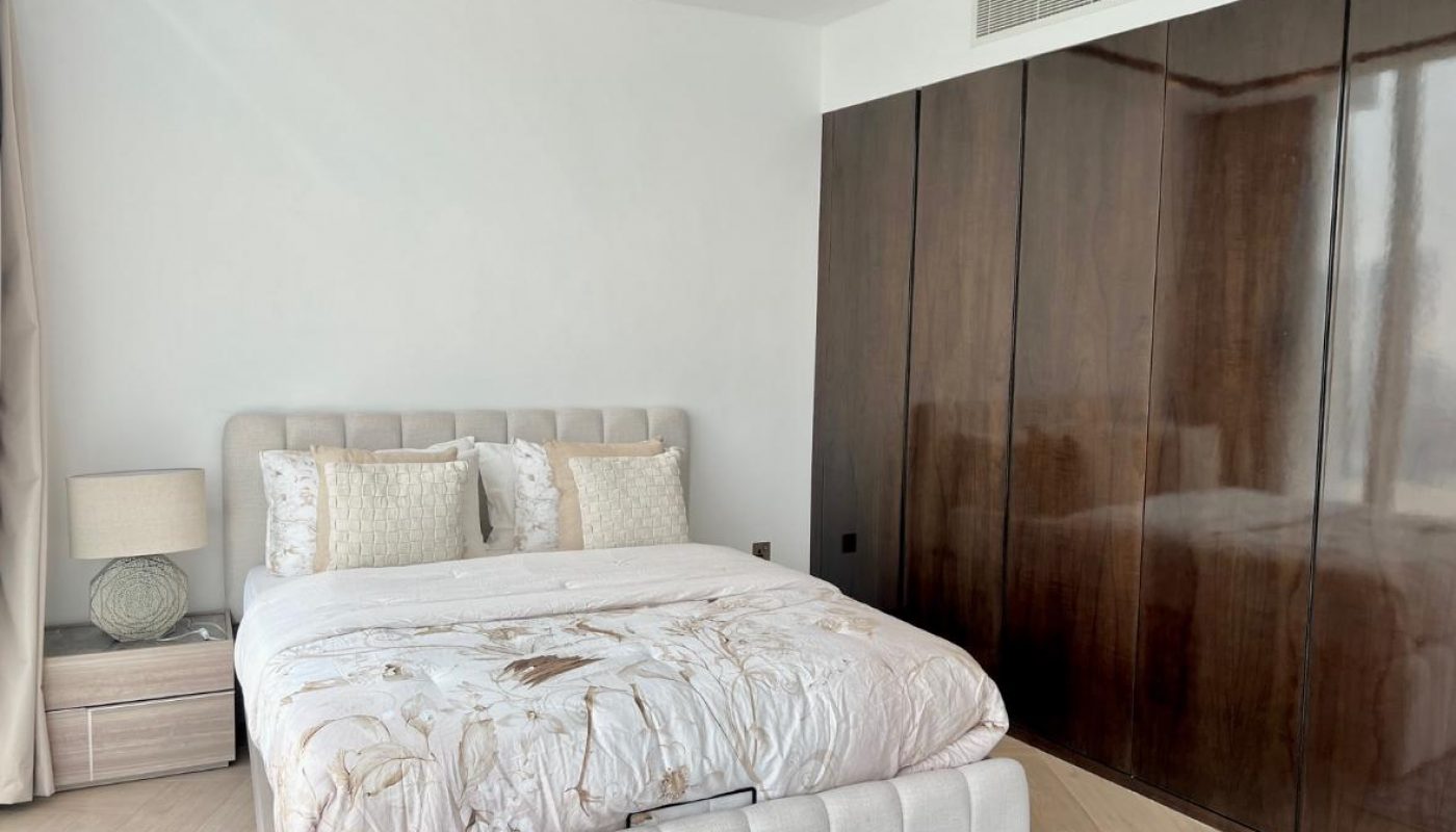 A modern bedroom with a beige upholstered bed, patterned bedding, a bedside table with a lamp, and large glossy brown wardrobes.