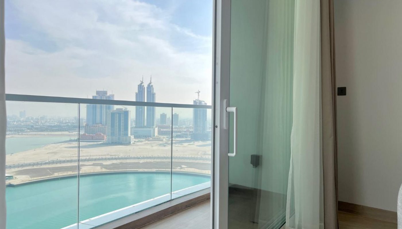 A glass door opens to a balcony with a view of high-rise buildings, a sandy area, and a body of water under a cloudy sky.