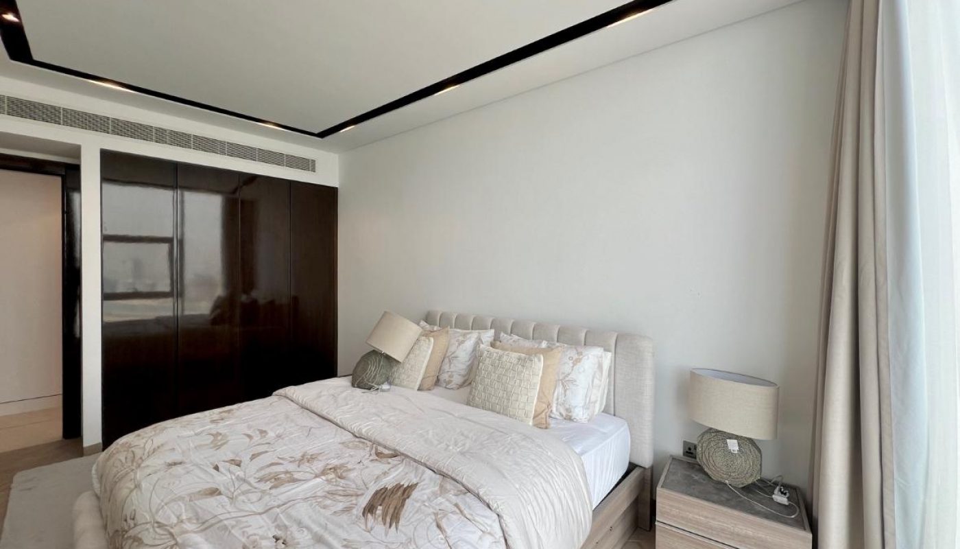 A modern bedroom with a large bed, decorative pillows, wooden nightstand, lamp, and floor-to-ceiling window with sheer curtains.