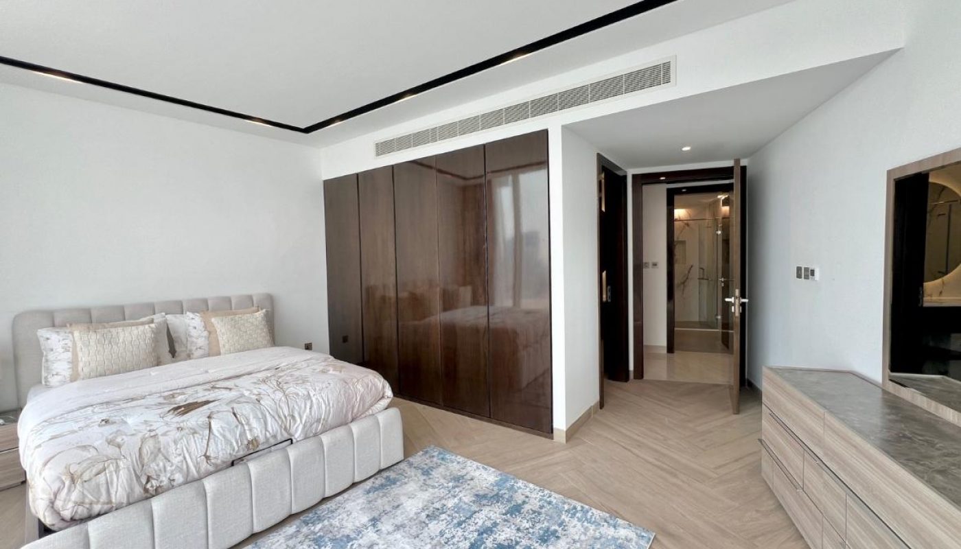 Modern bedroom with a large bed, wooden wardrobe, dresser, blue rug, and an open door leading to a bathroom. The room has neutral tones and natural lighting.