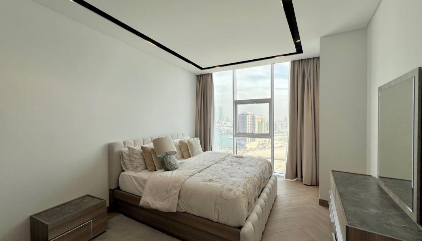 Modern bedroom with a large bed, two nightstands, a dresser with a mirror, light wood floors, and floor-to-ceiling windows showing a cityscape view.