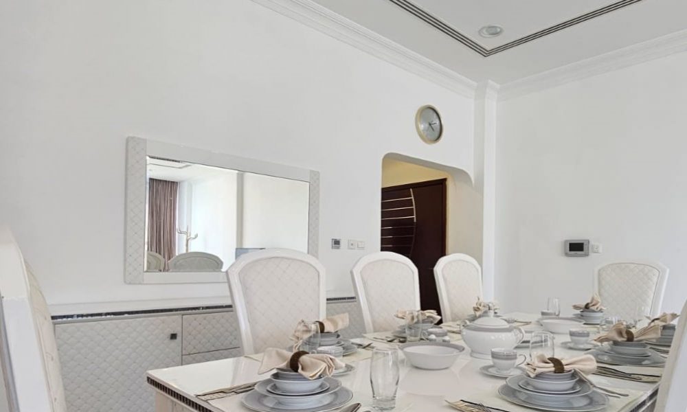 Elegant dining room with a white table set for six, featuring plates, bowls, utensils, and napkin holders. White cushioned chairs surround the table, and a large mirror hangs on the wall.