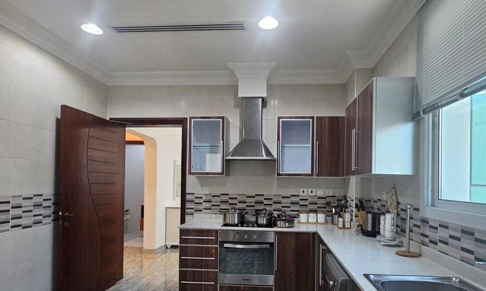 Modern kitchen with brown cabinets, stainless steel appliances, and white countertops. Tiled floor and walls, with a door leading to another room.