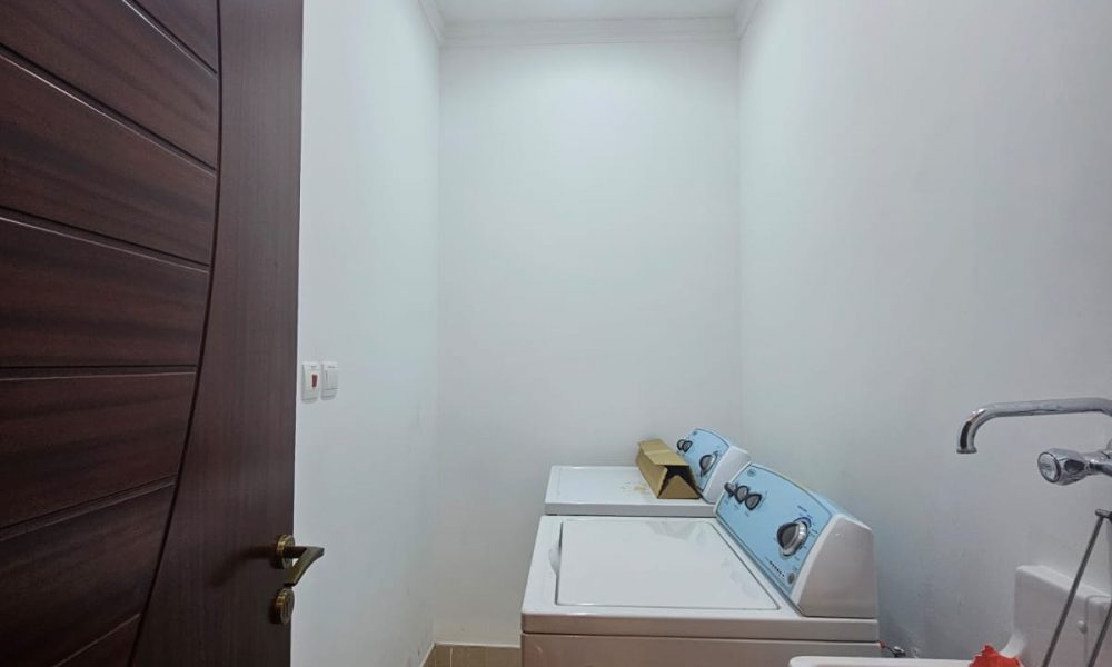 Laundry room with a wooden door, washing machine, dryer, and a sink with detergent bottles on the side. White walls and tiled floor.