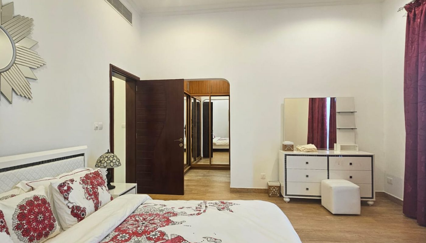 Bedroom with white and red floral bedding, a dresser with mirror, ottoman, and a rug. Two doors lead to other rooms, and the walls are decorated with framed art.