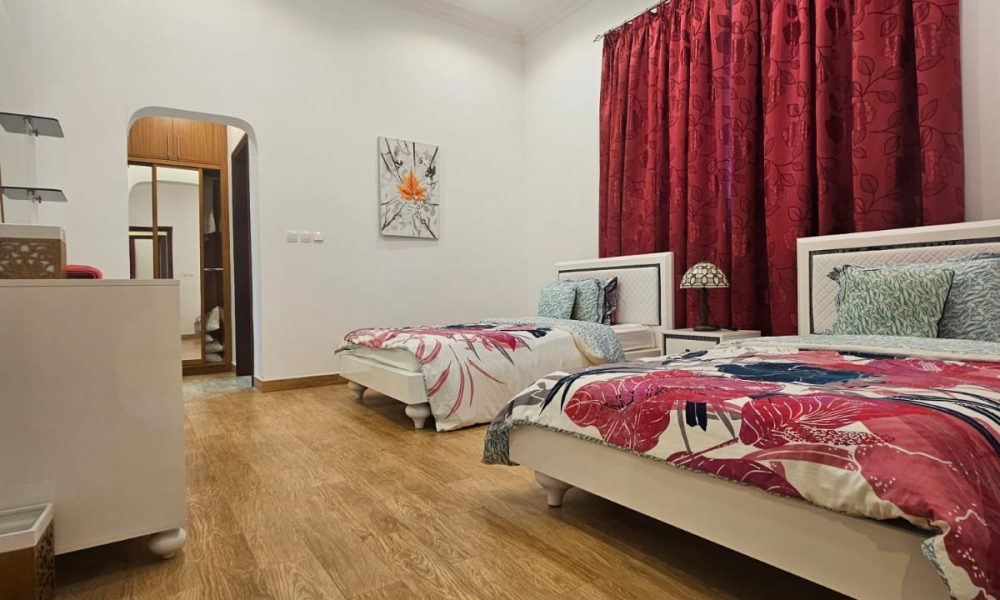 A bedroom with two single beds, colorful bedding, wooden floor, red curtains, a ceiling light, and a wall painting.