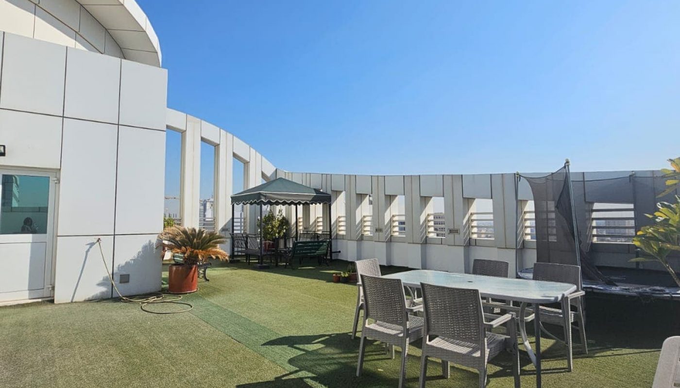 Rooftop terrace with artificial grass, a dining table with chairs, a tented seating area, and clear blue sky.
