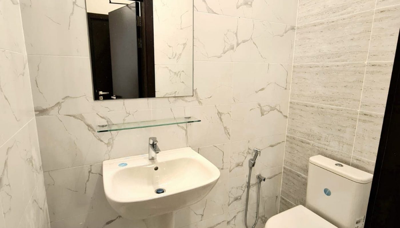 White bathroom with a marble-patterned tile design, featuring a wall-mounted sink, a mirror above, a toilet, and a handheld bidet spray on the side.