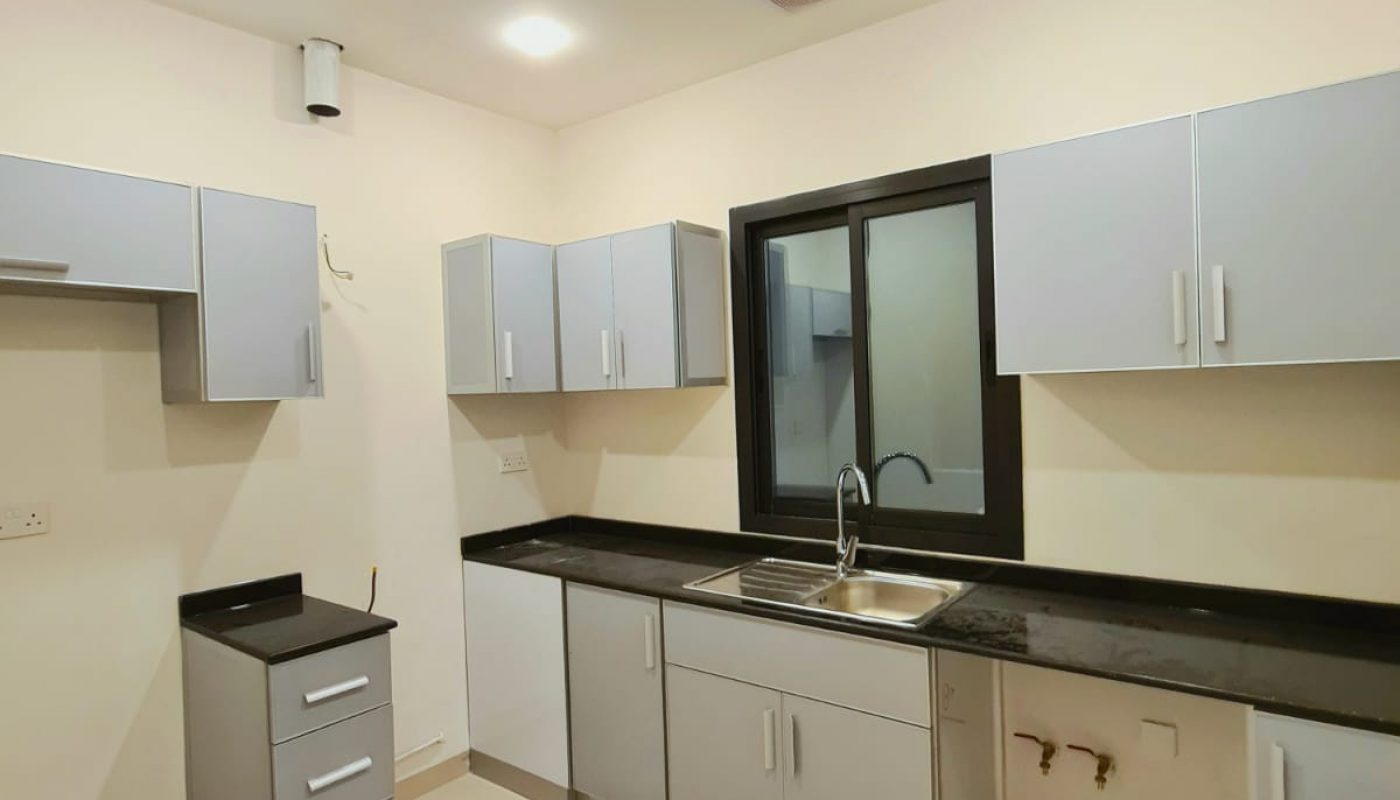 A modern kitchen with light gray cabinets, a black countertop, and a stainless steel sink. The floor is tiled, and the room is well-lit with recessed ceiling lights.