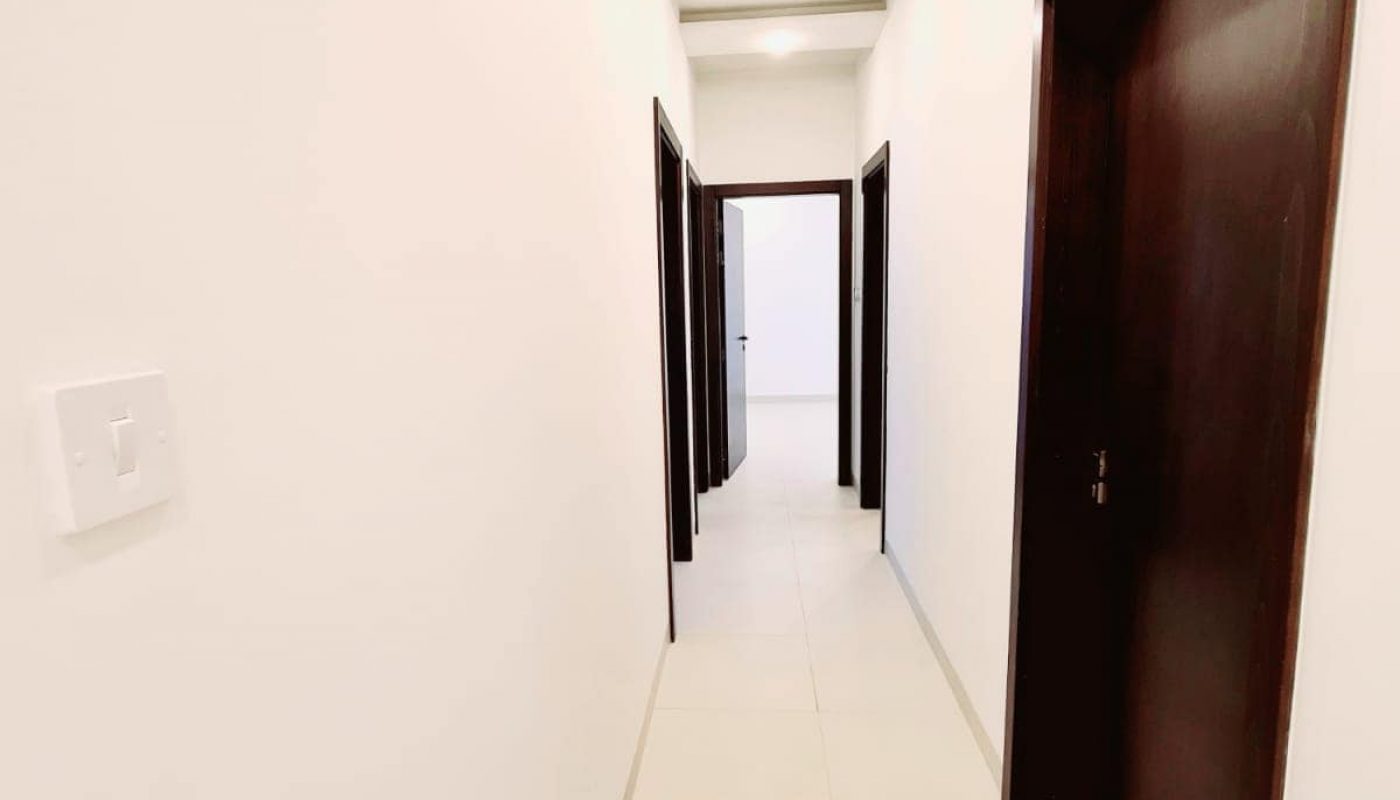 Long narrow hallway with white walls and tiled floor, leading to a series of dark wood doors on both sides. Ceiling features recessed lighting.