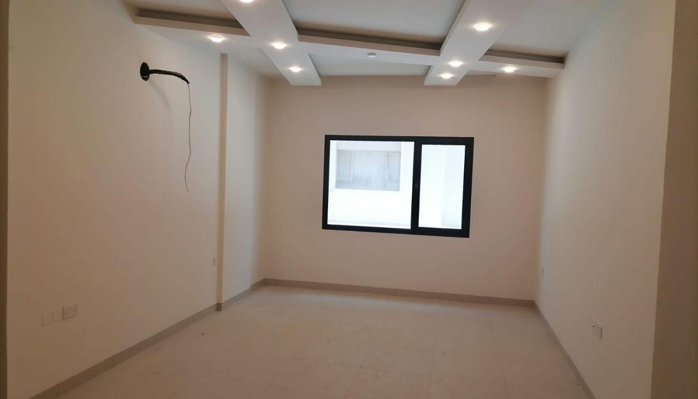 Empty room with a beige tiled floor, white walls, recessed ceiling lights, a wall-mounted TV bracket, and a window showing an exterior building.