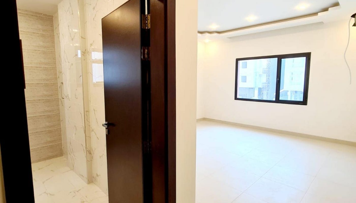 Minimalist interior with a partially open wooden door leading to a tiled room. The adjacent space features white walls, large windows, and recessed ceiling lights.