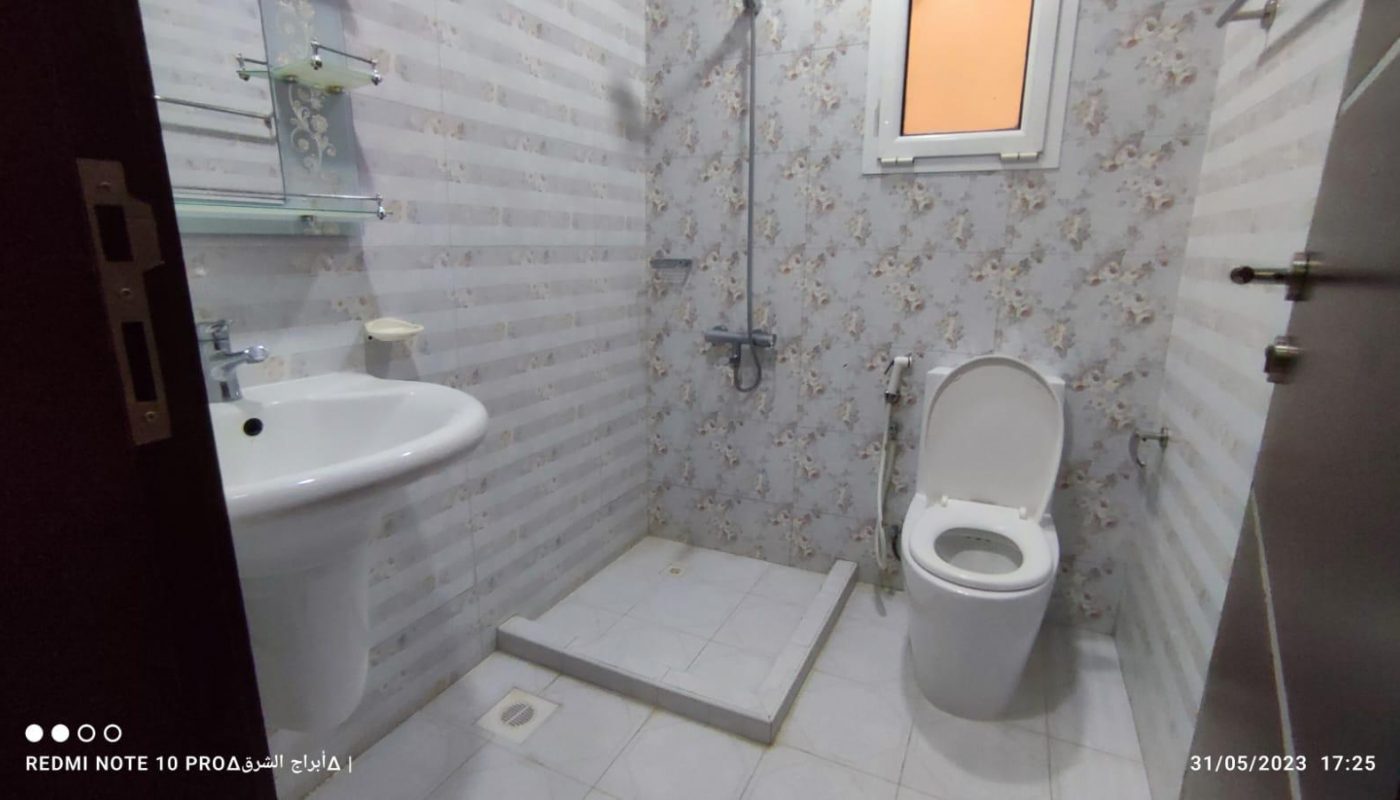 A bathroom with patterned walls, a sink, a toilet, and an open shower area. A small window is above the toilet, and the floor is tiled.