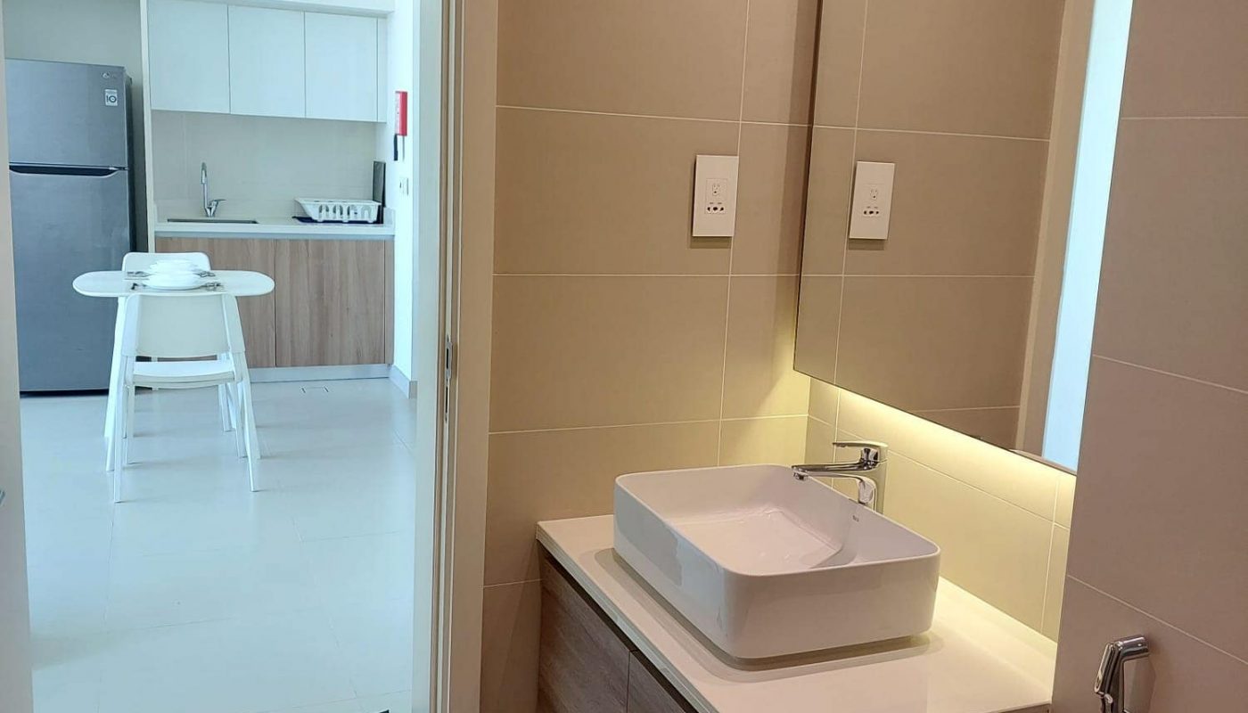 View from a bathroom with a sink and mirror, looking into a kitchen area with a refrigerator and a small table with stools.