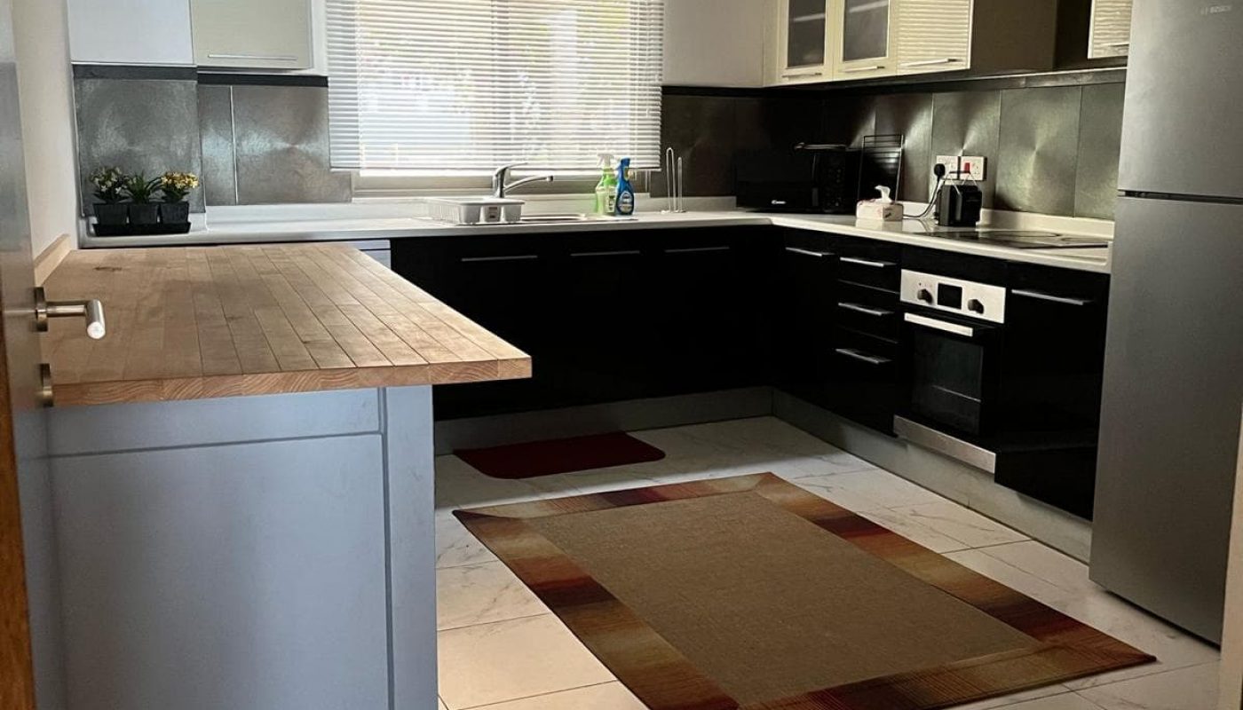 Modern kitchen with black cabinets, stainless steel appliances, wooden countertop, and a beige rug on white tile floor. A window with blinds is above the sink.