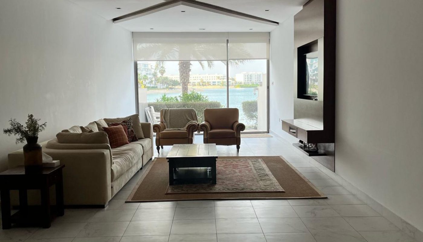 A modern, minimalistic living room with a sofa, armchairs, a coffee table on a rug, and a wall-mounted TV. Large sliding glass doors open to a view of trees and the outdoors.