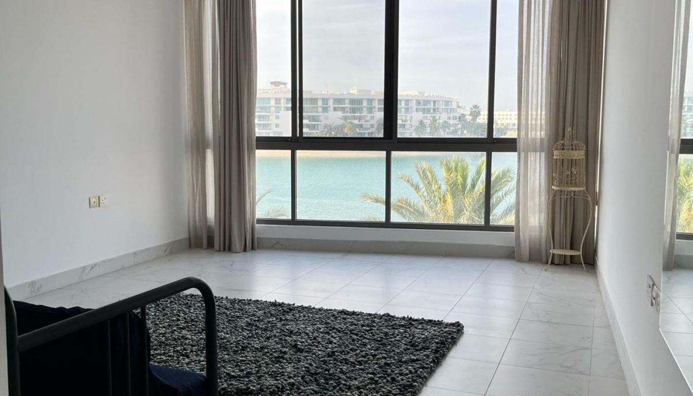 Minimalist room with large window overlooking water, white walls, a dark chair, a textured rug, and a hanging lamp.