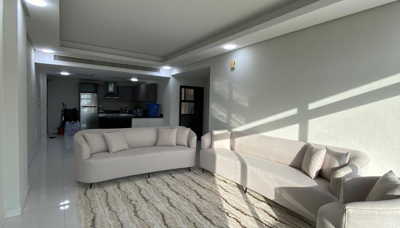 A modern living room with light-colored sofas, an armchair, a patterned rug, and an open view into the kitchen. Sunlight casts shadows across the floor and furniture.
