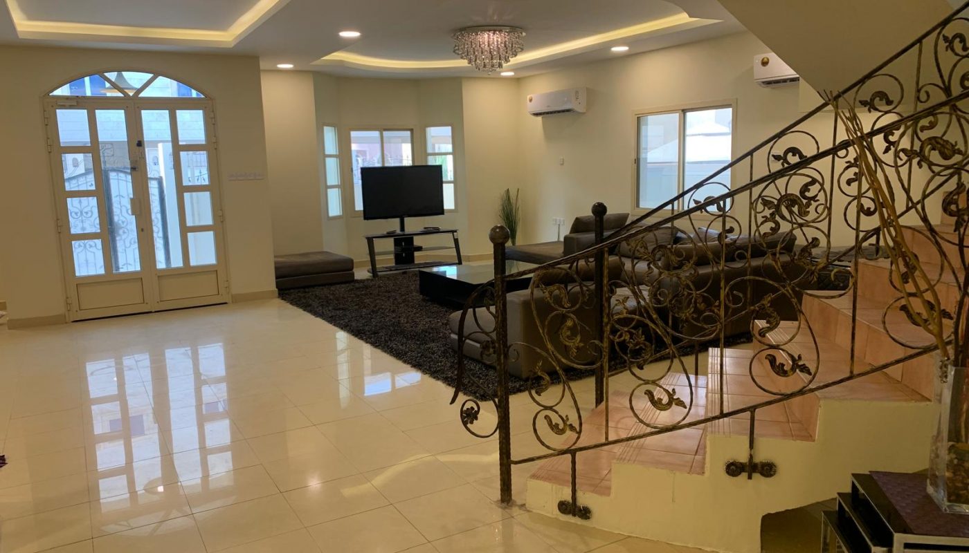 Spacious living room with tiled floor, brown sectional sofa, black rug, TV on stand, chandelier, and decorative iron staircase railing. Multiple windows provide natural light.