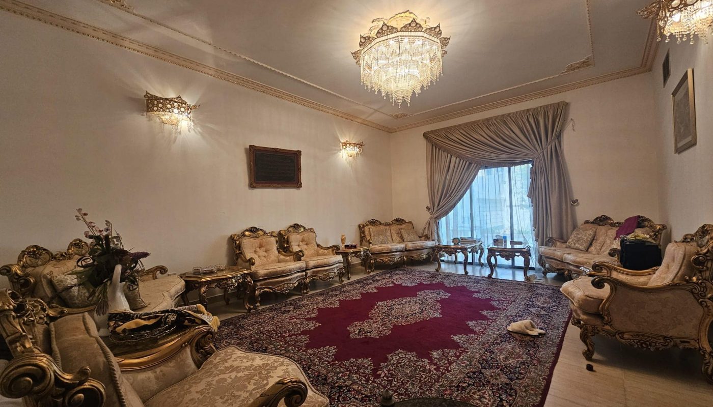 A luxurious living room with ornate gold-accented furniture, a large Persian rug, chandeliers, and a curtain-covered window.