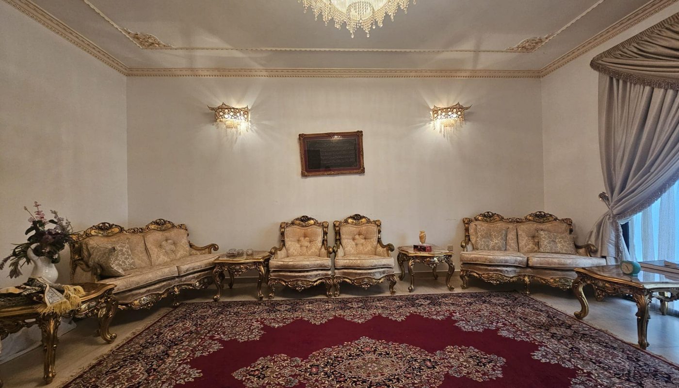 Elegant living room with a red patterned rug, ornate gold and cream furniture, a chandelier, wall lights, and draped curtains. A small side table with a vase and decor is on the left.