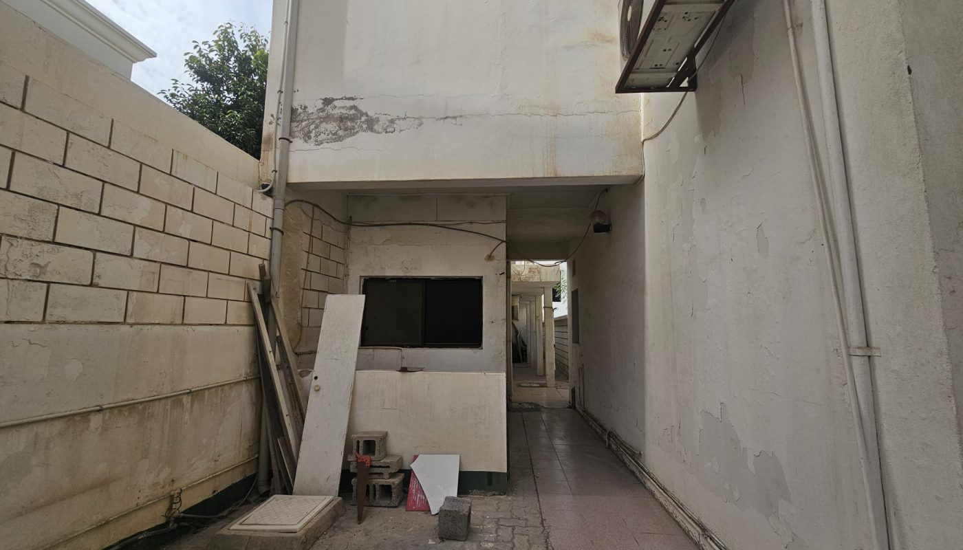 Narrow alley between two buildings with a tiled floor, an air conditioning unit, and construction materials leaning against a wall.