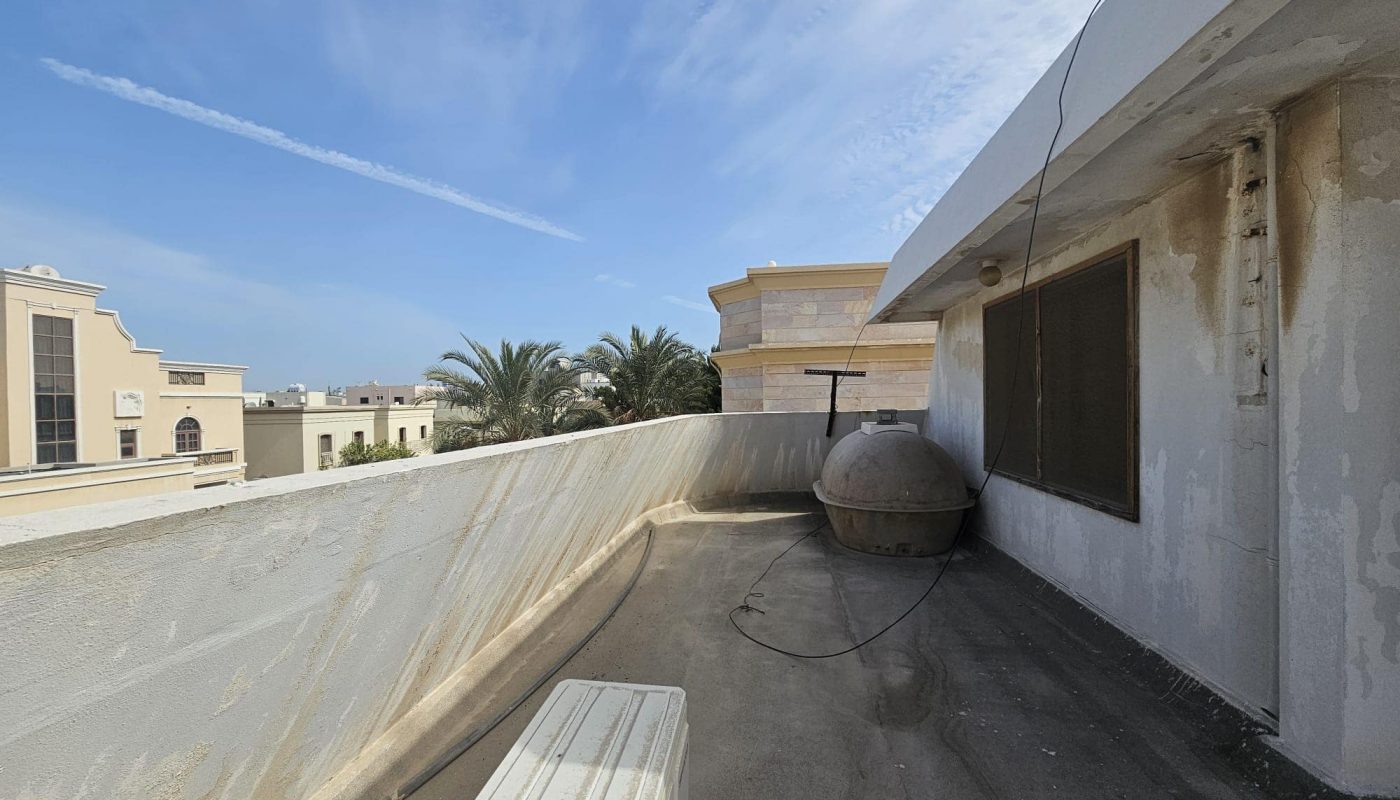Rooftop scene with a weathered water tank, concrete surface, and buildings in the background. Palm trees and a clear sky are visible.