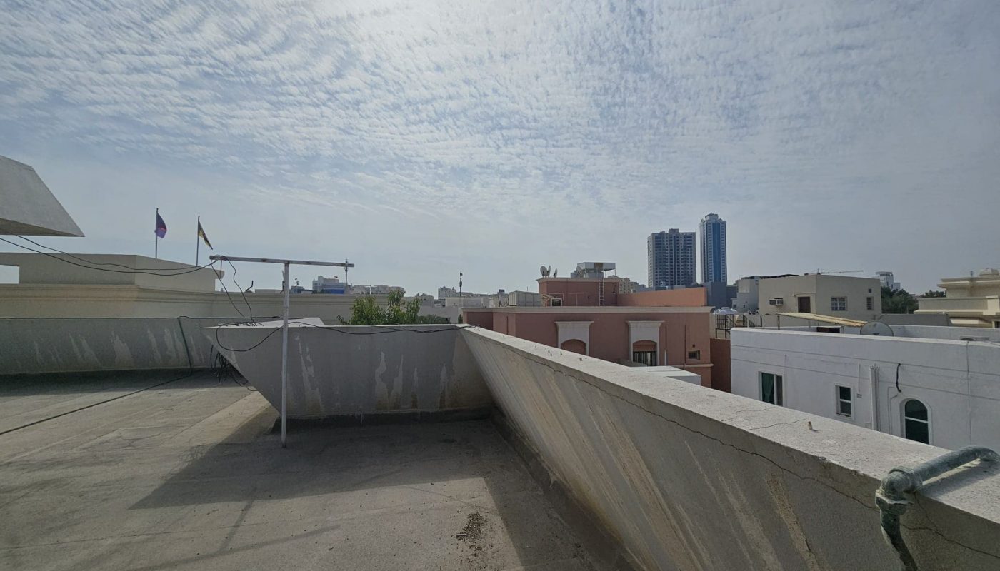 Rooftop view of a residential area showing buildings, a clear sky with scattered clouds, and two tall towers in the distance.