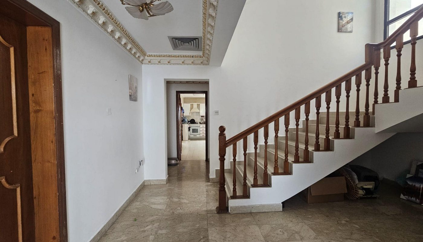 Inside a house featuring a marble floor, wooden staircase, and decorative ceiling. A hallway leads to a kitchen area.